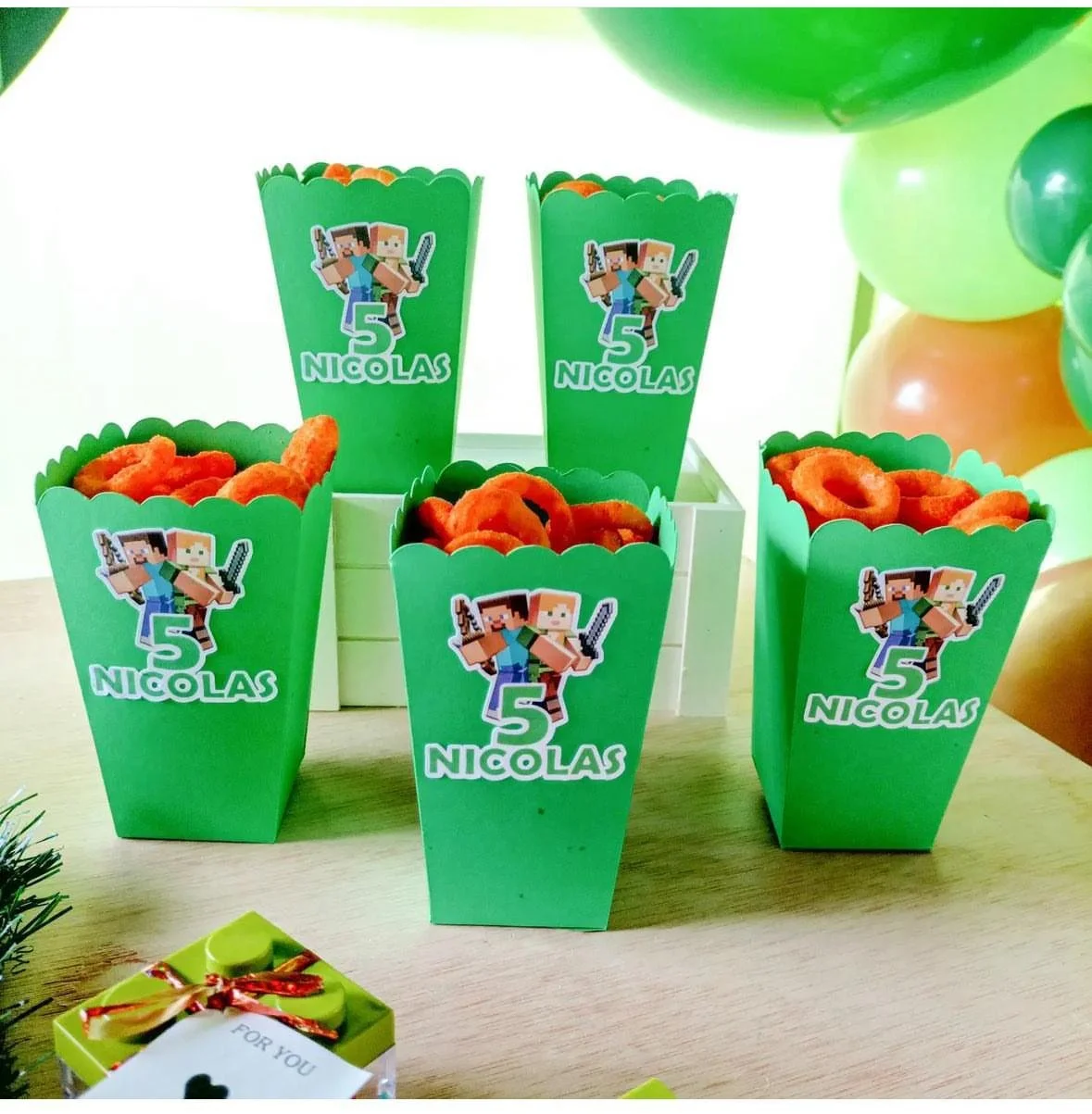Contenitori verdi a forma di popcorn in cartone con decorazione di Minecraft, numerati 5 e con il nome Nicolas, contenenti snack a forma di anello. Sul tavolo, in primo piano, ci sono piccoli regali incartati colorati. In background palloncini verdi,