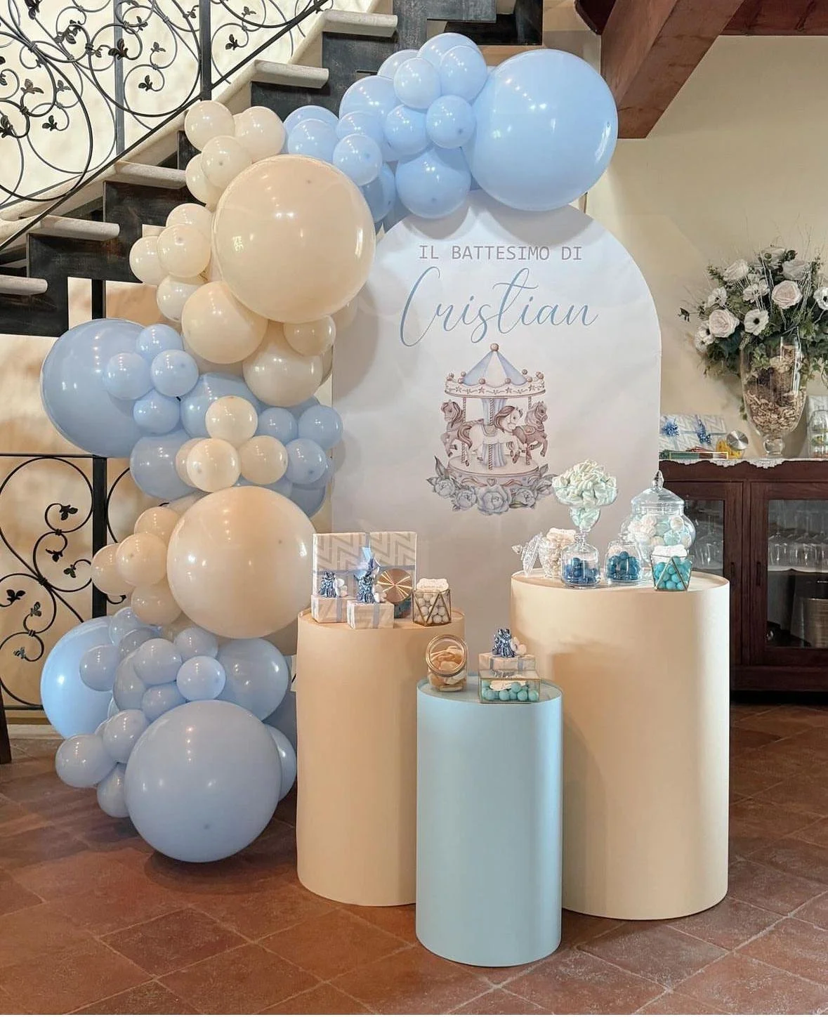 Decorazione per battesimo con palloncini in colori blu e crema, una targa con scritto 'Il battesimo di Cristian' e un carosello eccentrico come tema, con tavoli decorativi con regali e dolci.