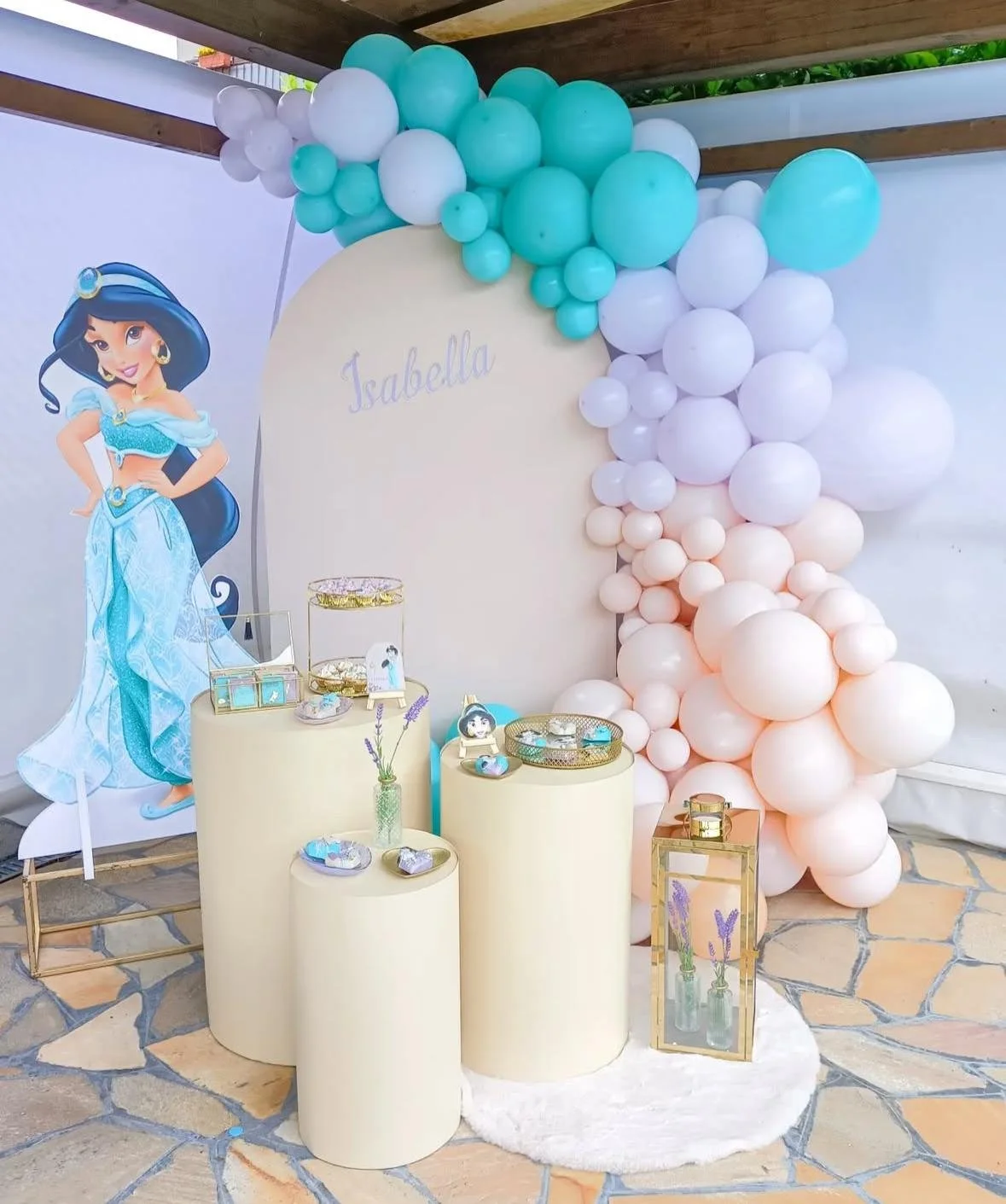 Decorazione per festa di compleanno con poster di Jasmine, palloncini pastello in toni azzurro, bianco e rosa, tavolo con decorazioni e piccoli regali. La parola "Isabella" scritta sulla parete