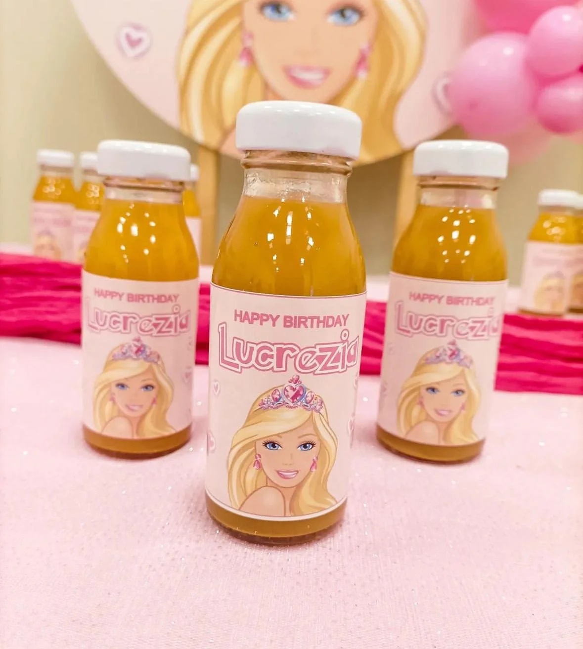 Mini bottiglie di succo con etichetta a tema Barbie, decorazioni rosa e un poster di una Barbie sullo sfondo in occasione di un compleanno.