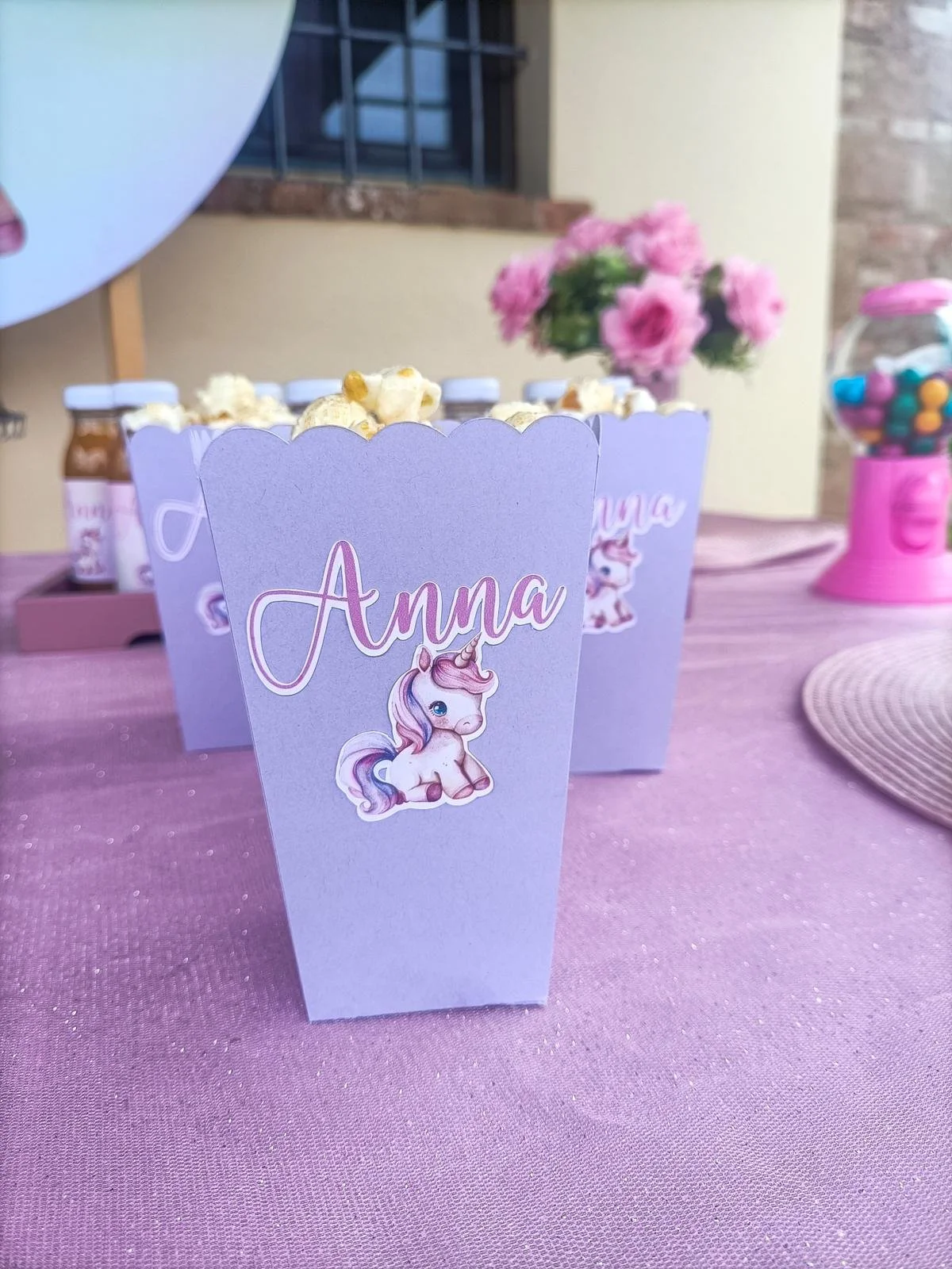 Decorazione per festa di compleanno con targhe rosa con un unicorno e il nome Anna, su tavolo con tovaglia rosa, in uno spazio all'aperto. Inoltre, ci sono bottigliette e un distributore di caramelle rosa.