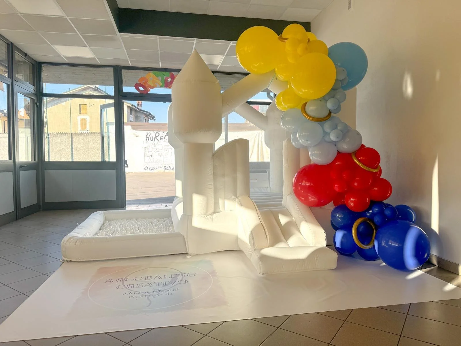 Una mongolfiera gonfiabile bianca decorata con palloncini colorati gialli, rossi, blu e grigi, posizionata all'interno di una stanza con pareti di vetro e un pavimento di piastrelle.