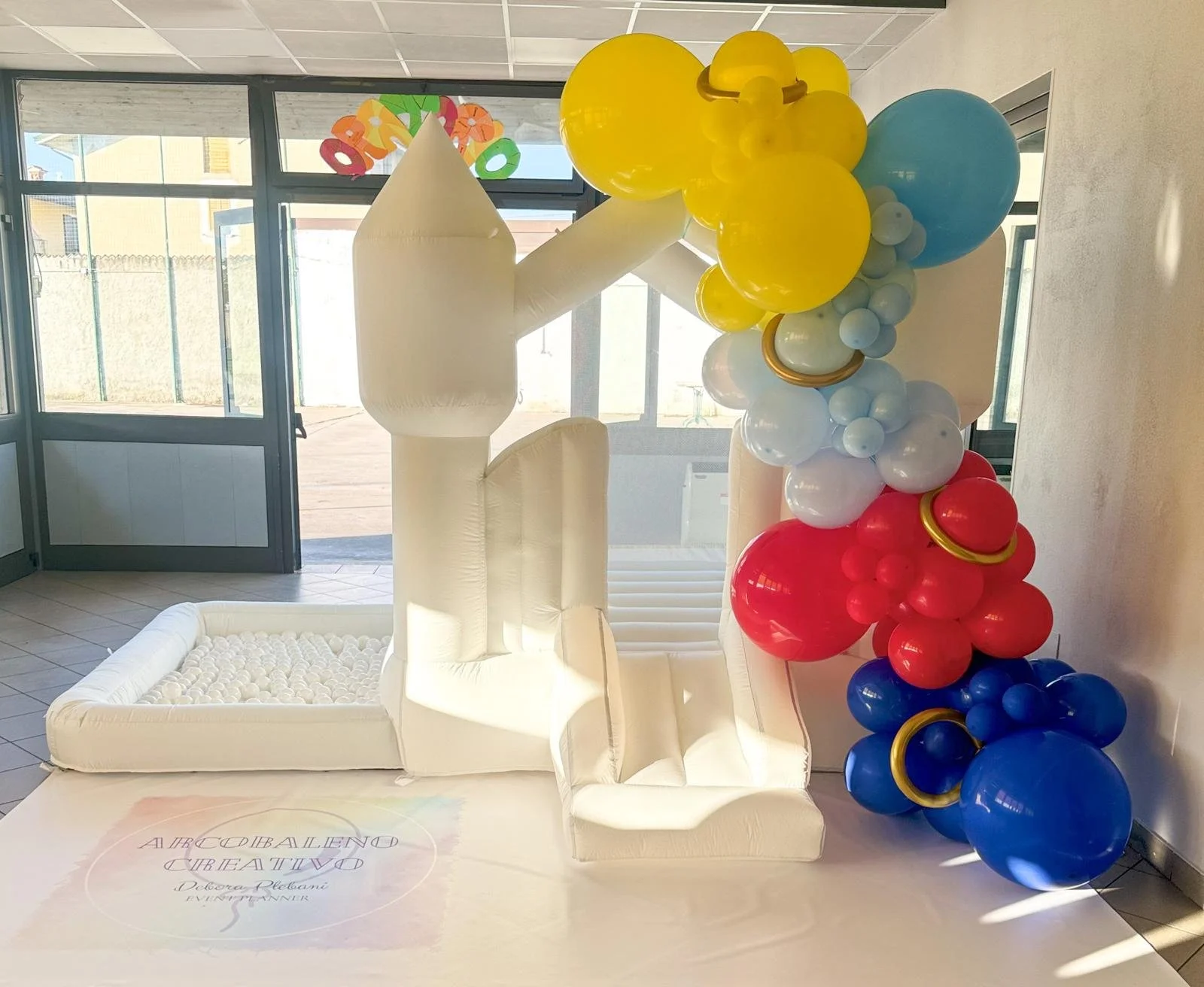 Decorazione con gonfiabili a forma di letto con palline bianche, con un grande arco di palloncini colorati in rosso, giallo, bianco e blu accanto.