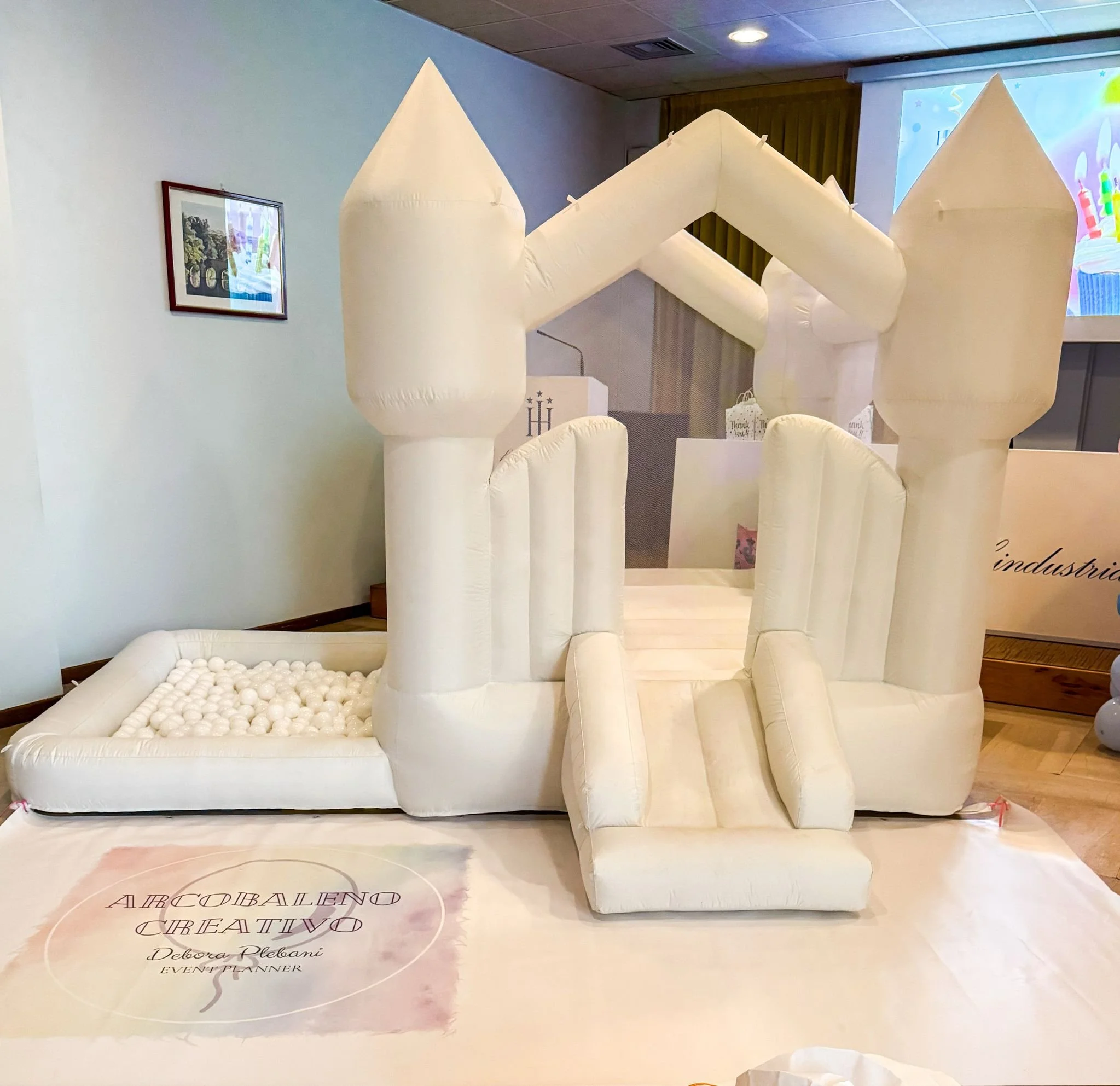 Gioco gonfiabile a forma di castello bianco in stile fiabesco con torri e scaletta, posizionato su un pavimento in legno in una sala decorata per un evento.
