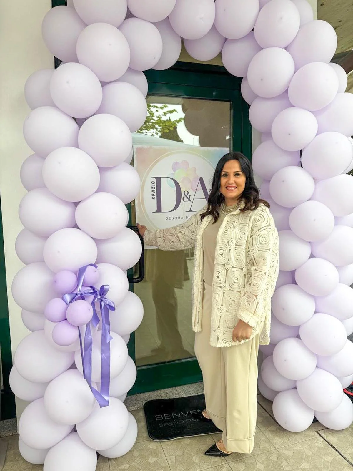 Una donna sorridente davanti a un ingresso decorato con archi di palloncini bianchi e lilla, che tiene una mano sulla porta.