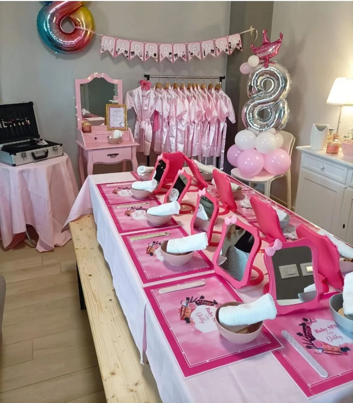 Decoração rosa per un baby shower con tavolo preparato con federe, specchi, tazzine, asciugamani e cartelloni con tema femminile e baby shower.