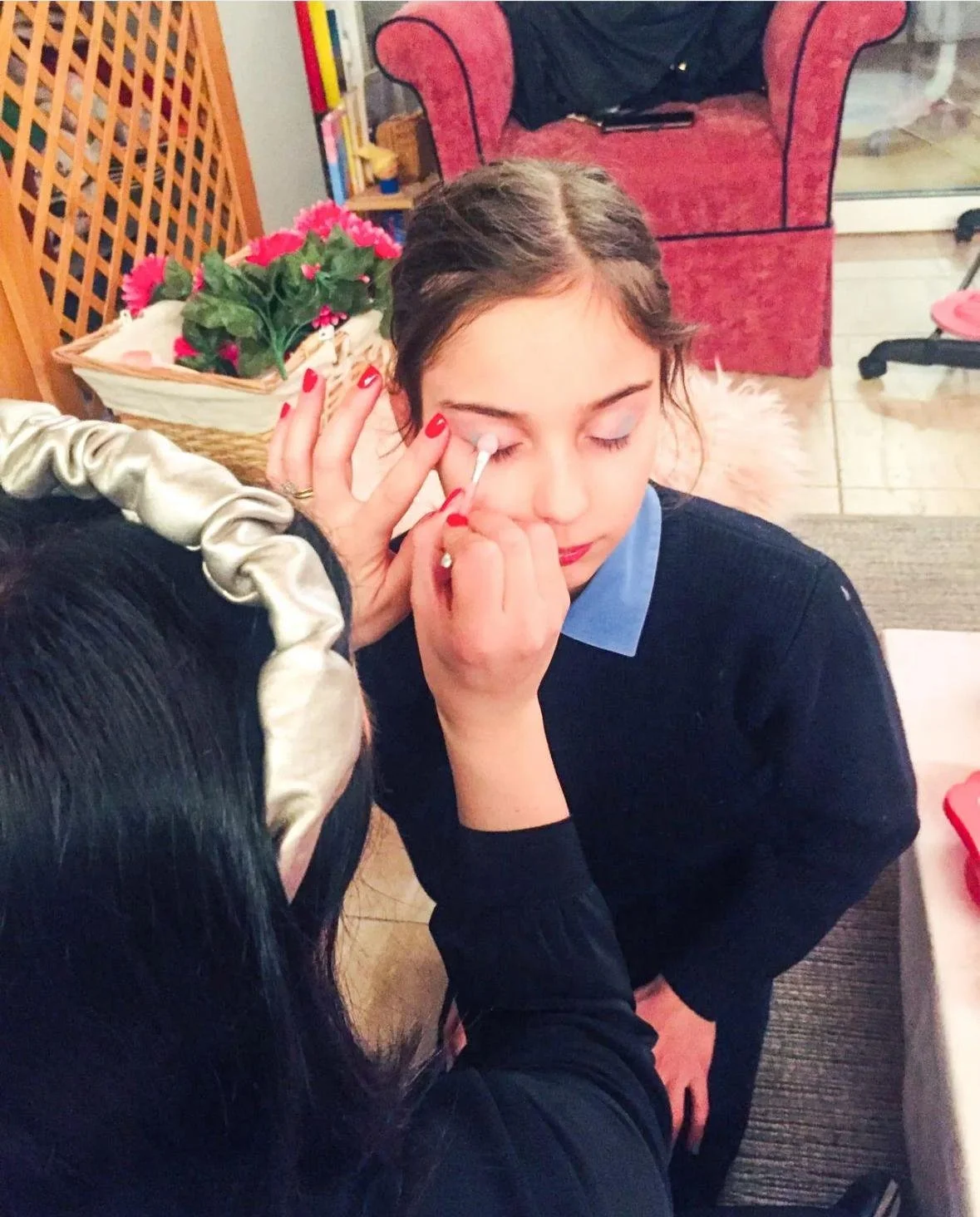 Una donna riceve un trucco sugli occhi da un'altra persona. La donna ha i capelli castani, indossa una camicia azzurra e un maglione nero. La scena si svolge in un ambiente domestico con arredi come una poltrona rossa, un cesto di fiori rosa e altri 