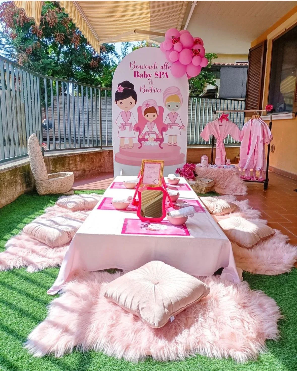 Decorazione per una festa di baby spa con un tavolo rotondo coperto da una tovaglia bianca, cuscini e tappeti rosa e pelosi, nello sfondo un grande cartello con illustrazioni di bambine in accappatoio e testo "Benvenuti alla Baby SPA di Beatrice". A 