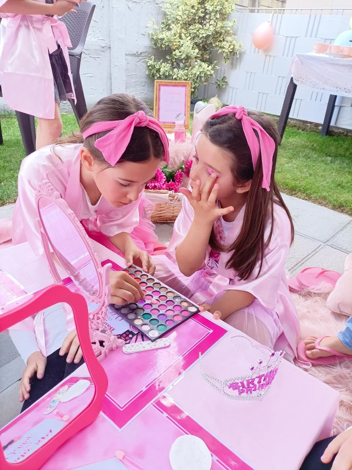 Due bambine truccando una l'altra durante una festa di compleanno all'aperto, con decorazioni rosa e un cappello di coroncina sulla tavola.