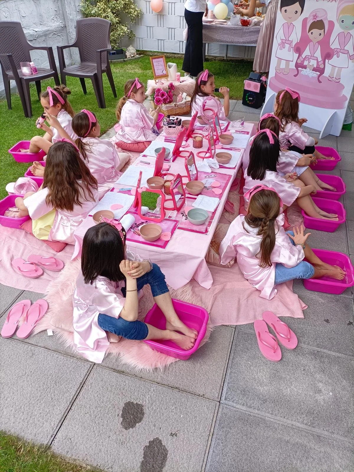 Bambine sedute su un tappeto rosa, in abiti rosa, partecipano a una festa in stile tea party su un tavolo decorato con oggetti rosa e specchi, con ciabatte rosa ai piedi e all'aperto.