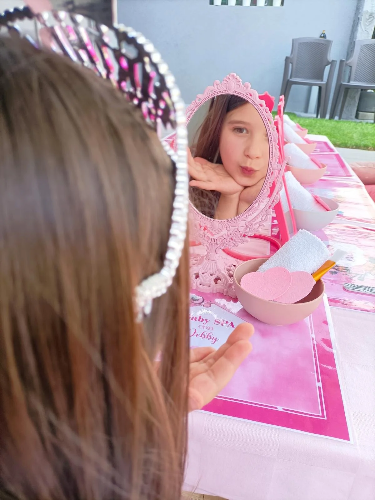 Una bambina con una fascia decorativa si guarda allo specchio; la scena è decorata con rosa e contiene accessori per il taglio di capelli o il trucco, tra cui un tovagliolo e una spatola.