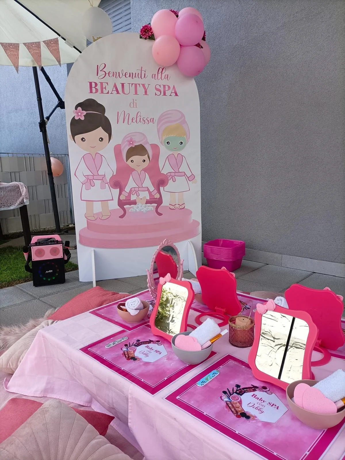 Decorazione a tema spa di bellezza per bambina con tavolo rosa, specchietti, candele e decorazioni rosa, e un cartellone con illustrazioni di bambole in stile cartoon.