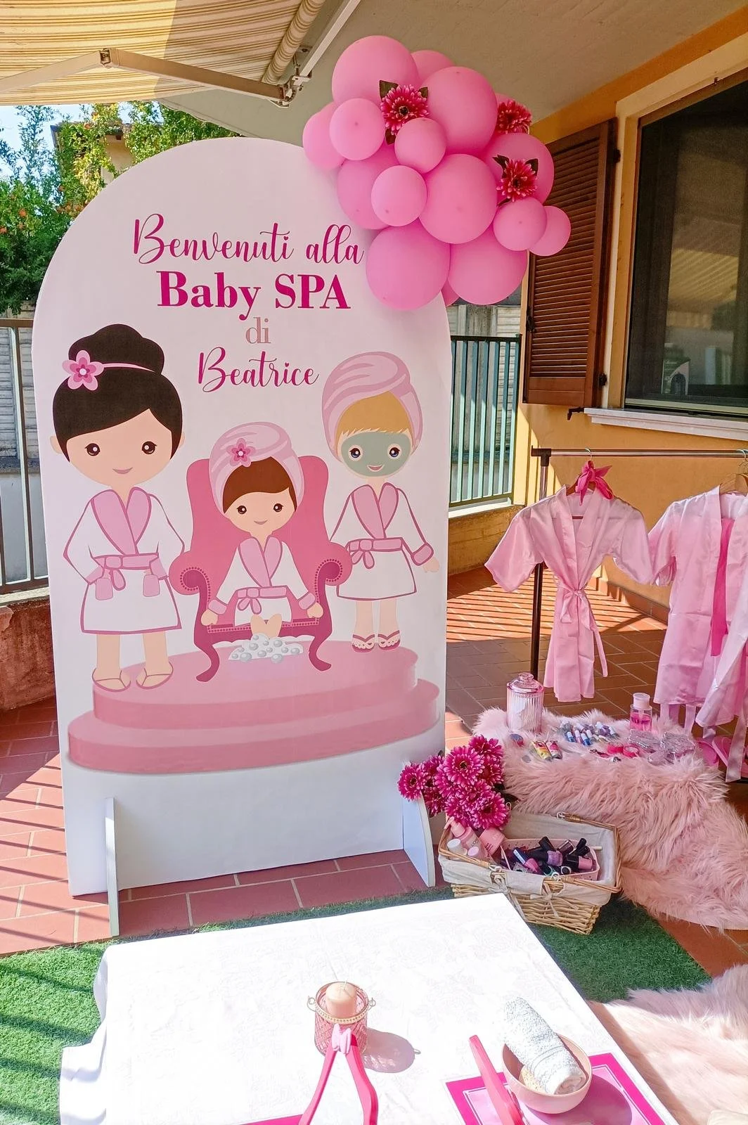 Decorazione rosa per festa di benvenuto alla Baby Spa di Beatrice, con immagini di bambole stilizzate, palloncini rosa e un'area con vestitini rosa, oggetti per manicure e fiori rosa.