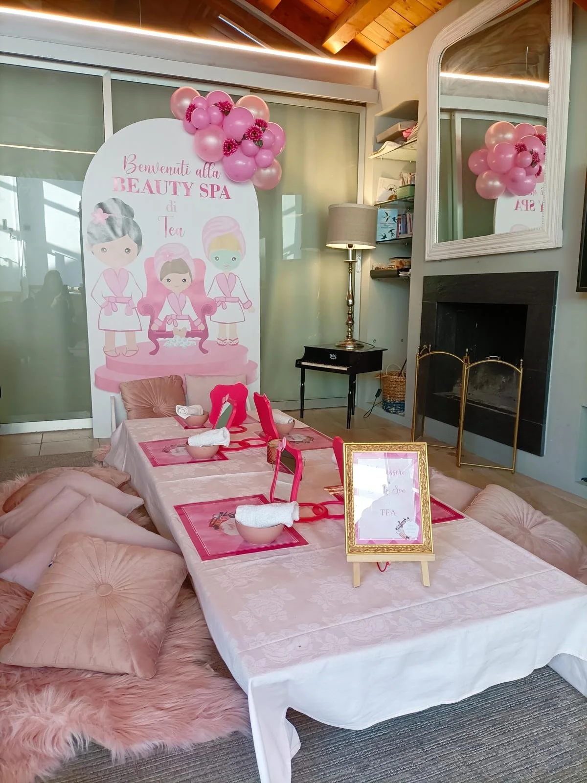 Decorazioni per una spa di bellezza a tema rosa con palloncini, figura di tre bambole in accappatoio, e un tavolo apparecchiato con tovagliette rosa e asciugamani arrotolati in ciotole. Murale con scritta in italiano, grande specchio, e camino