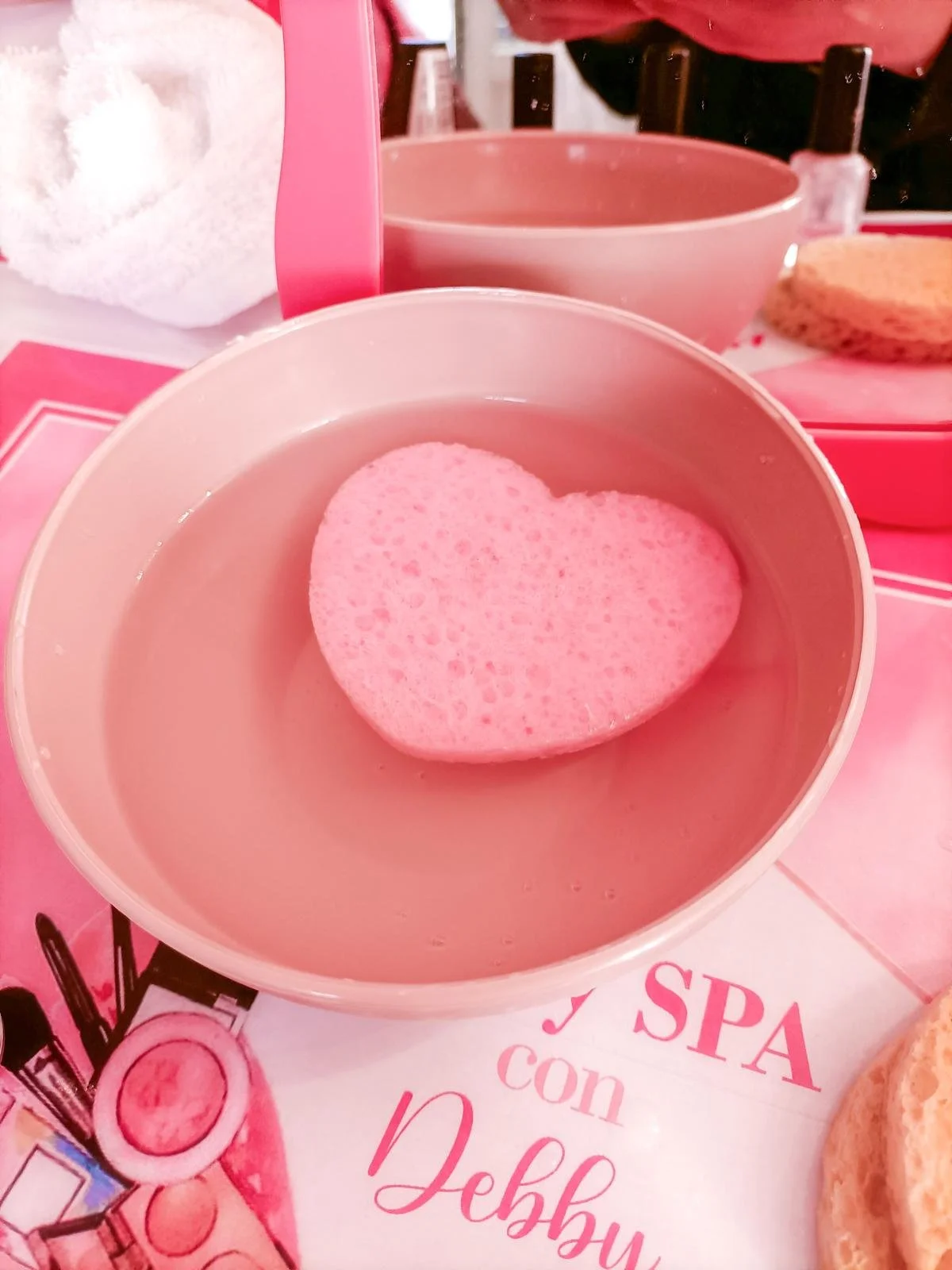 Un cuore di spugna rosa galleggia in una ciotola con acqua, su un tavolino rosa con scritte, in un ambiente tipicamente spa o bellezza.