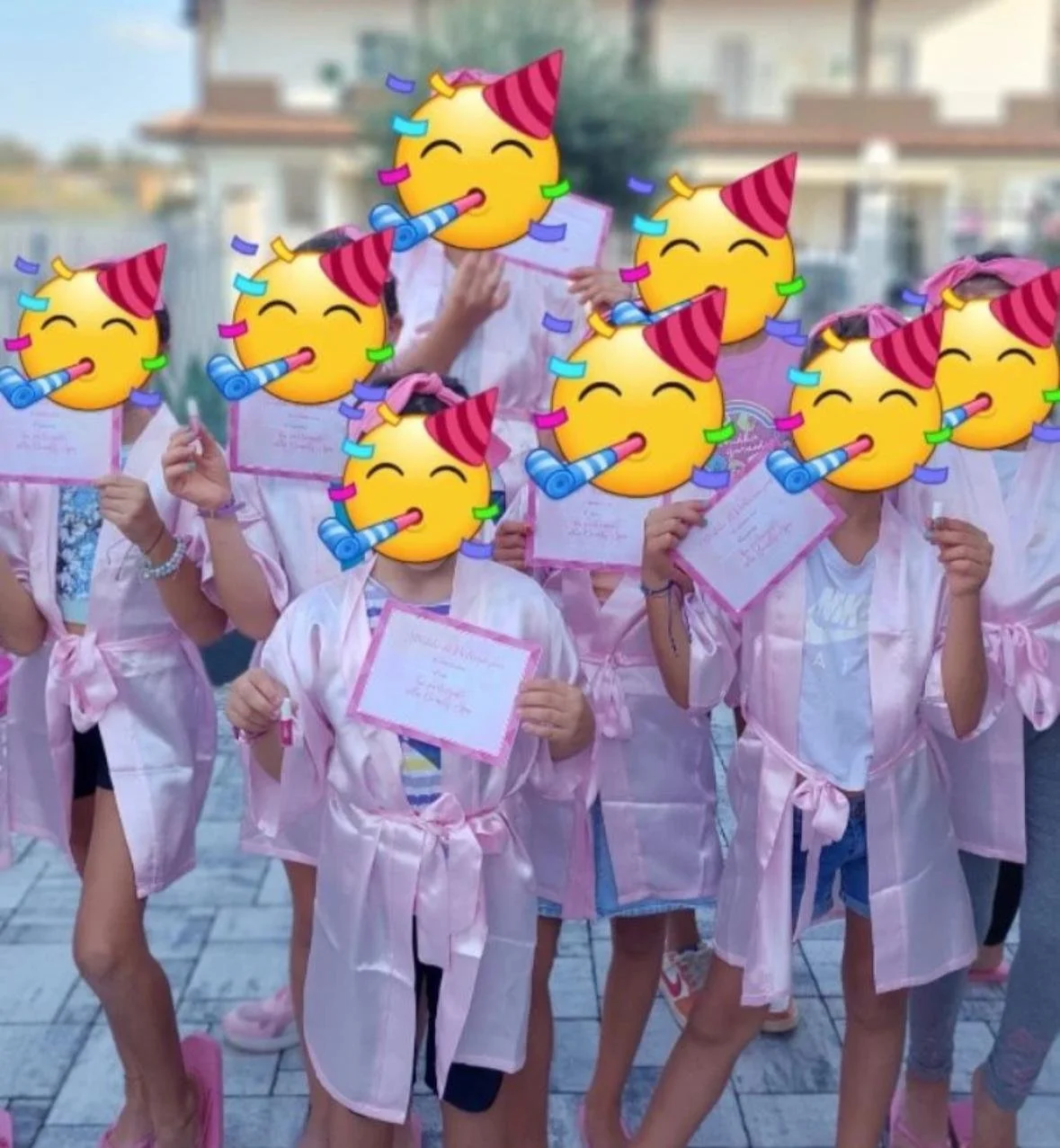 Gruppo di persone vestite in pigiama rosa, con emoji di faccine con Cappello di Festa e corno di festa che coprono i volti, che tengono attestati e brindano con bicchieri, in un evento festivo esterno.