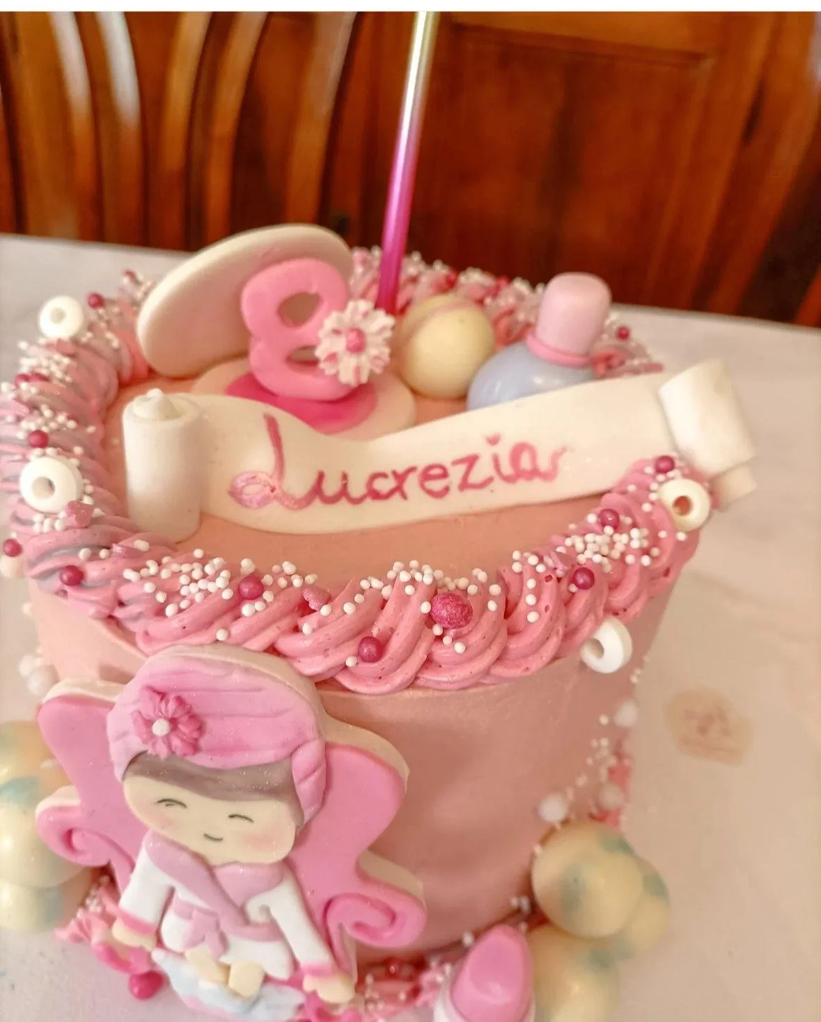 Torta decorata con tema magico rosa e bianco, con figure di una fata e decorazioni di zucchero, con la scritta 'Lucrezia'.