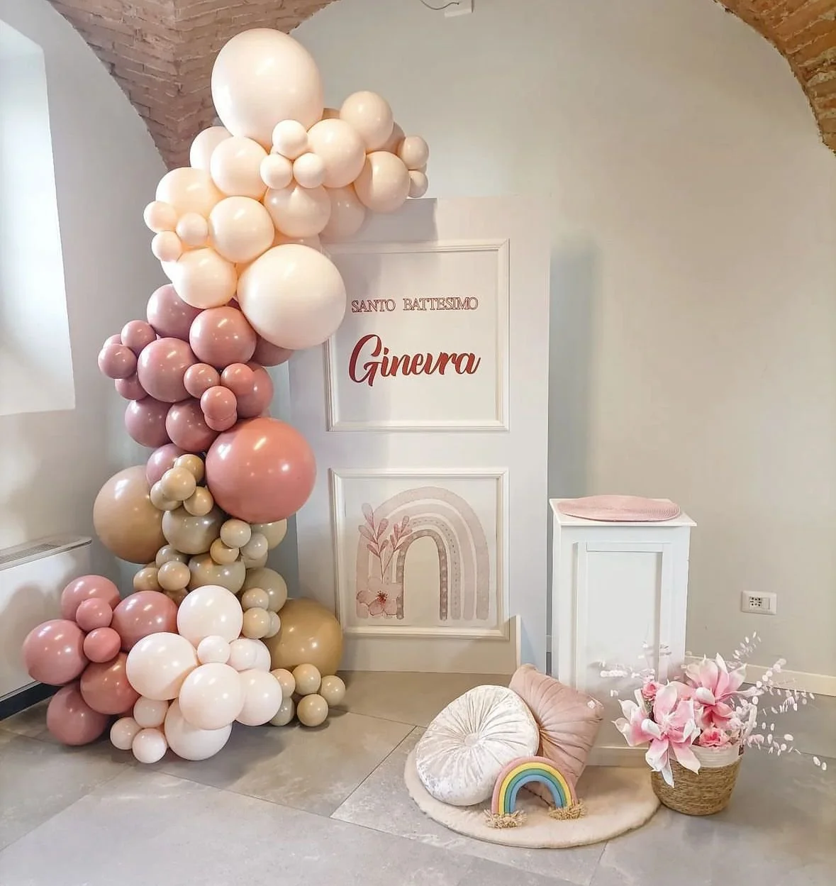 Decorazioni per battesimo con palloncini rosa e crema, quadro con scritta 'Santo Battesimo Gineura', fiori rosa, cuscini e arco decorativo.