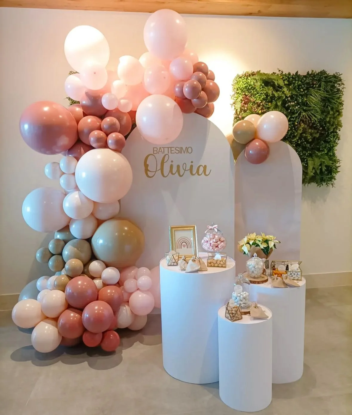 Decorazione per battesimo con palloncini rosa, bianco, beige e marroni, accompagnata da un tavolo con regali e fiori, con sfondo di pannelli decorativi e parete verde