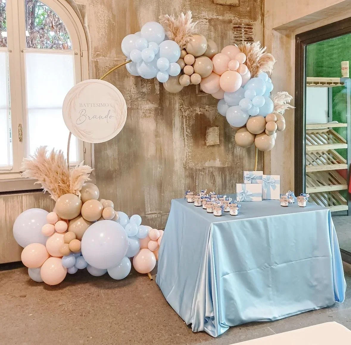 Decorazione per battesimo con un arco di palloncini in colori pastello, una tavola coperta con tessuto azzurro e regalini posizionati sopra, in una stanza con pareti in cemento e finestre con tende bianche.