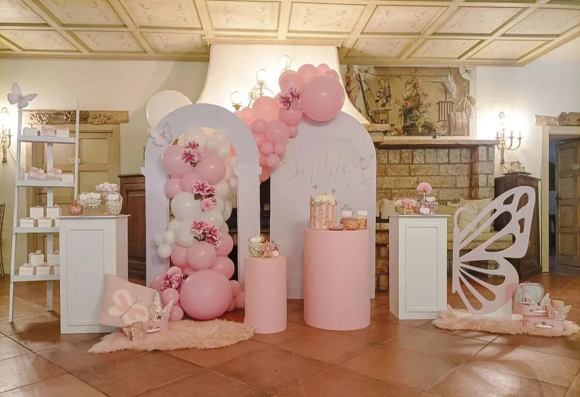Decorazioni per una festa di compleanno a tema farfalla, con palloncini rosa e bianchi, addobbi floreali, strutture di cartone a forma di farfalla, e una torta decorata in stile rosa, ambientate in una stanza elegante con camino in pietra.