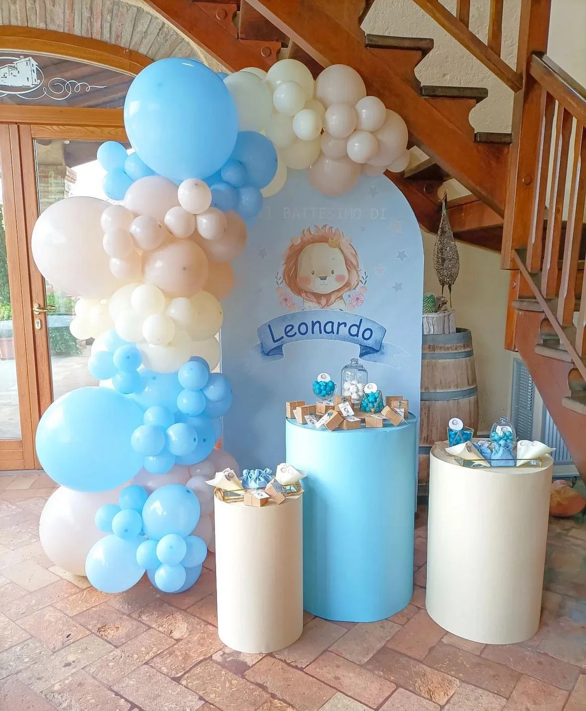 Decorazione per battesimo con palloncini bianchi e azzurri, un backdrop con una testa di leone e il nome Leonardo, tavoli con confetti e regali, una cascata di palloncini e un menu decorativo.