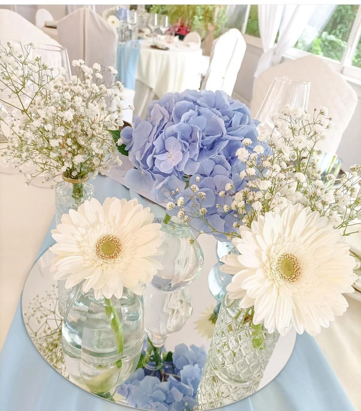 Centro tavola con fiori di ortensia blu, gerbere bianche e fiori di baby breath in vari vasi di vetro su un piatto rotondo riflettente, decorato con petali di ortensia blu.
