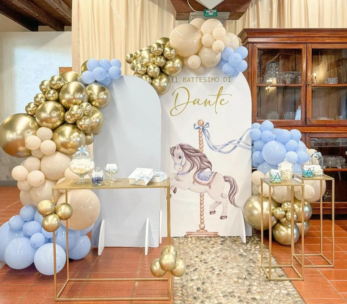 Decorazione per battesimo con palloncini color oro e azzurro, sfondo con immagine di cavallo a dondolo e scritta in italiano 'Il battesimo di Dante'.