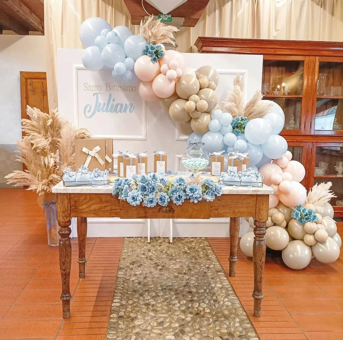 Decorazione per battesimo con palloncini pastello in toni azzurro, beige e rosa, fiori in azzurro, tavolo ricco di regali e dolci, sfondo con scritta "Santo Battesimo Julian"