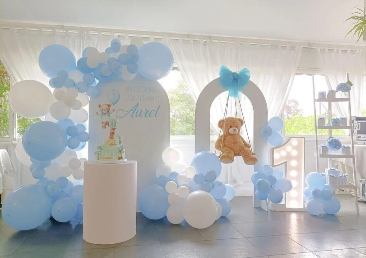 Decorazioni per festa di compleanno di Aurel, con palloncini bianchi e azzurri, un grande numero uno illuminato, un orsetto di peluche e un arco decorato con un fiocco azzurro.
