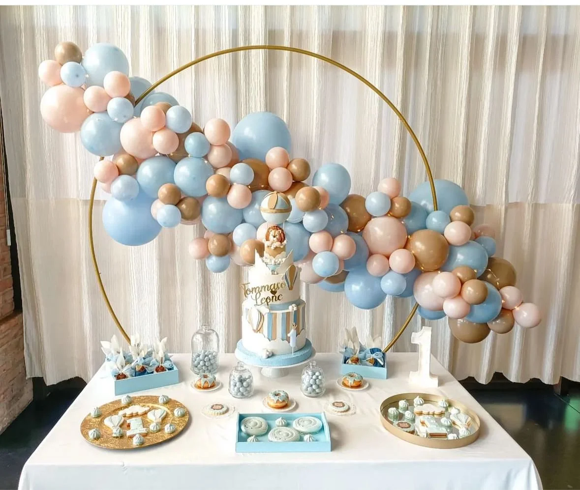 Tavolo decorato con dolci e un grande arco di palloncini di colori celeste, rosa e oro, con un'icona di un palloncino e una nuvole. Al centro una torta decorata con tema aerostatico, con un piccolo aereo e una nuvola, e una scritta in italiano.