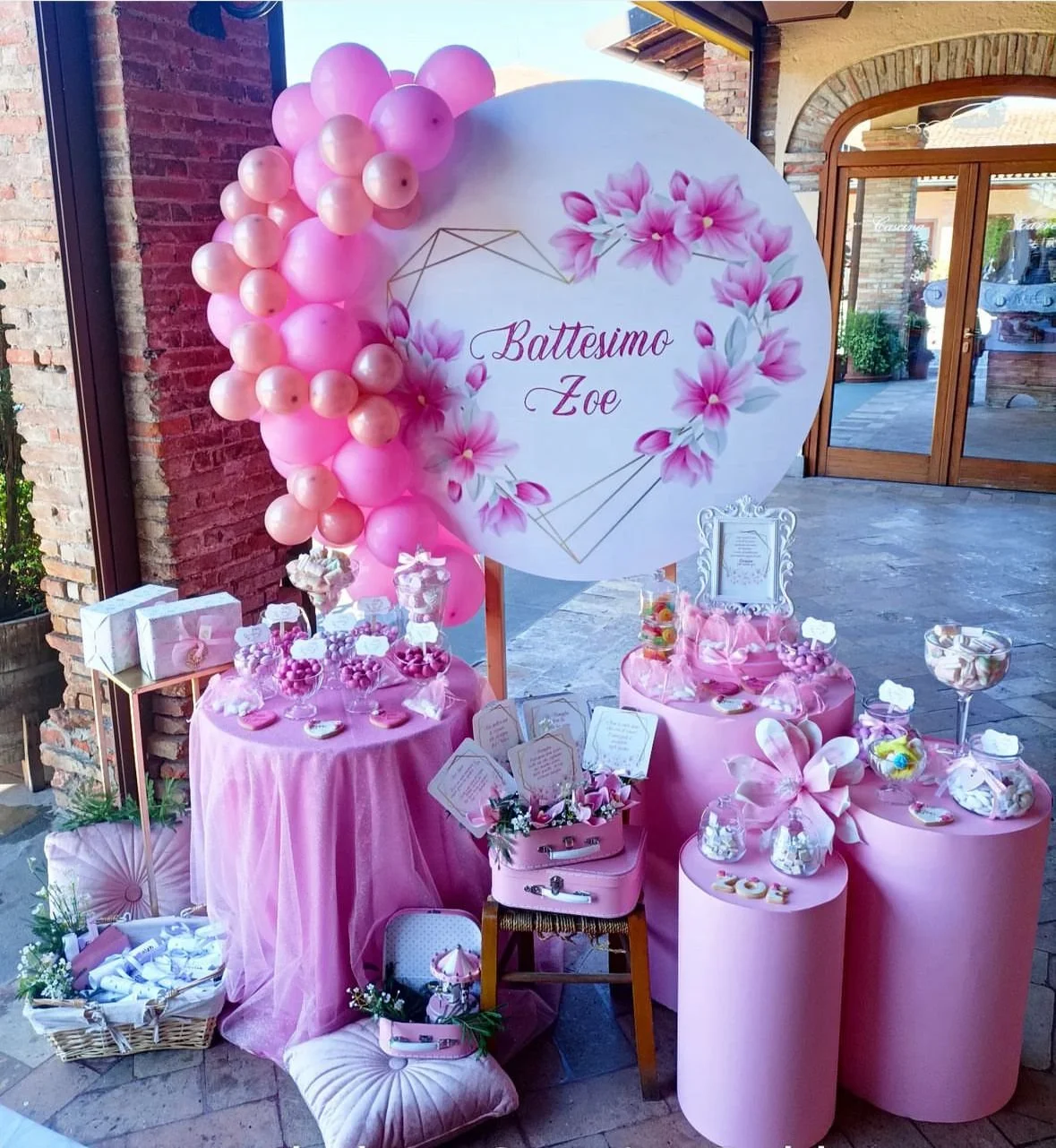 Decorazione per battesimo con fiori rosa, palloncini rosa e tavoli con dolci e regali, sullo sfondo segno con scritto 'Battesimo Zoe'.