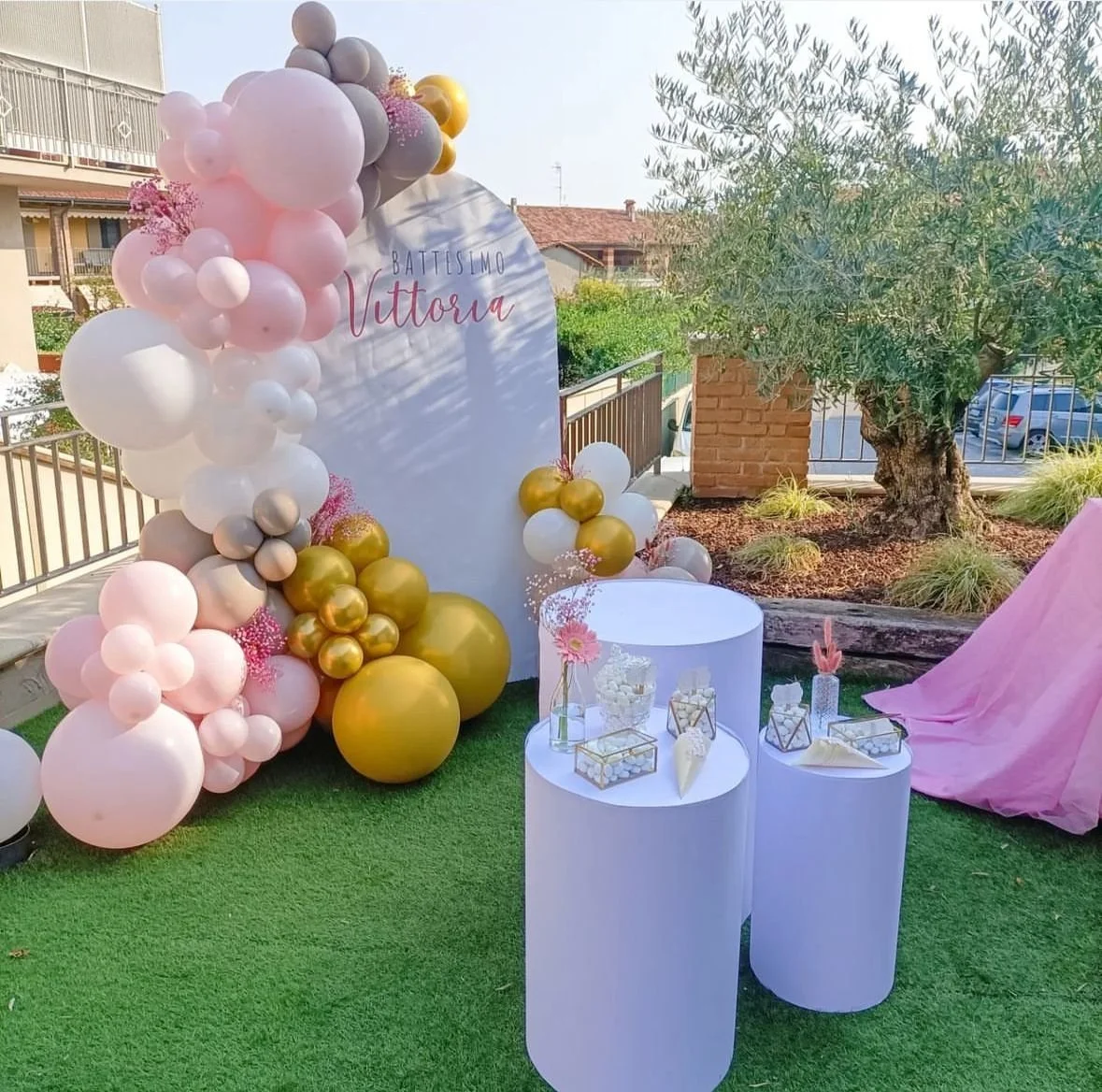 Setup decorativo con palloncini rosa, bianco, oro, e grigio per la celebrazione del battesimo di Vittoria, con un'area con tavolini bianchi, fiori e decorazioni.