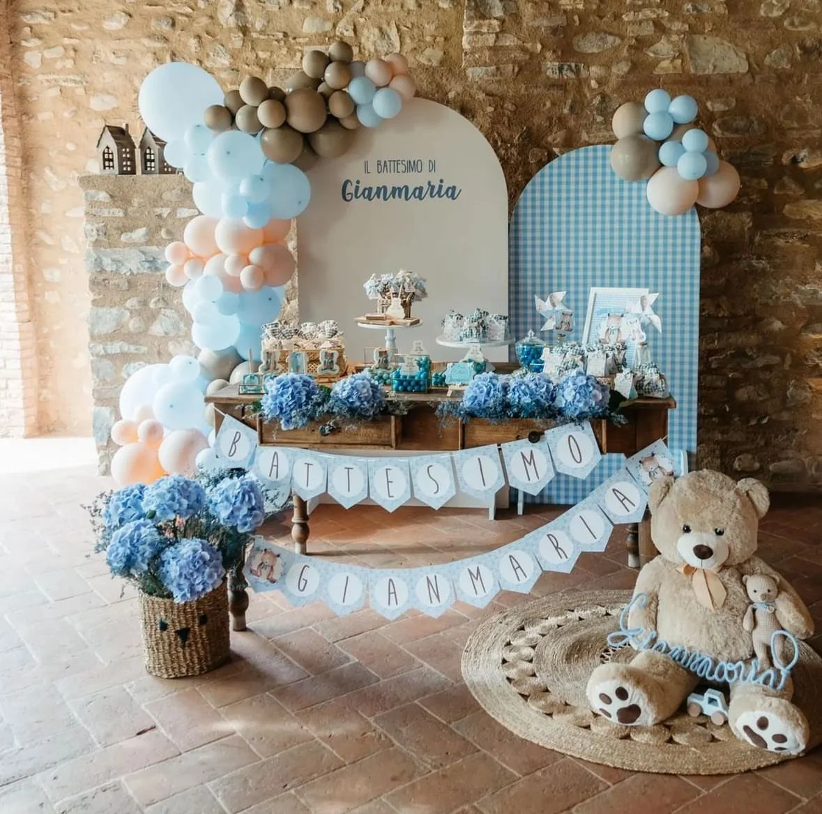Decorazione per battesimo di bambino con palloncini azzurri, fiori blu, teddy bear, e banner con scritte in italiano.