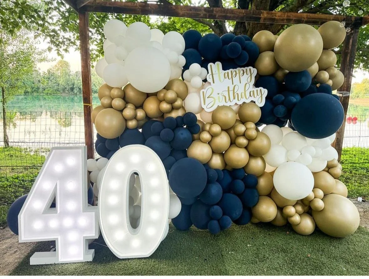 Decorazione di compleanno con palloncini nei colori bianco, oro e blu, con un grande numero 40 illuminato, un cartello con scritta 'Happy Birthday', e un pannello in legno.