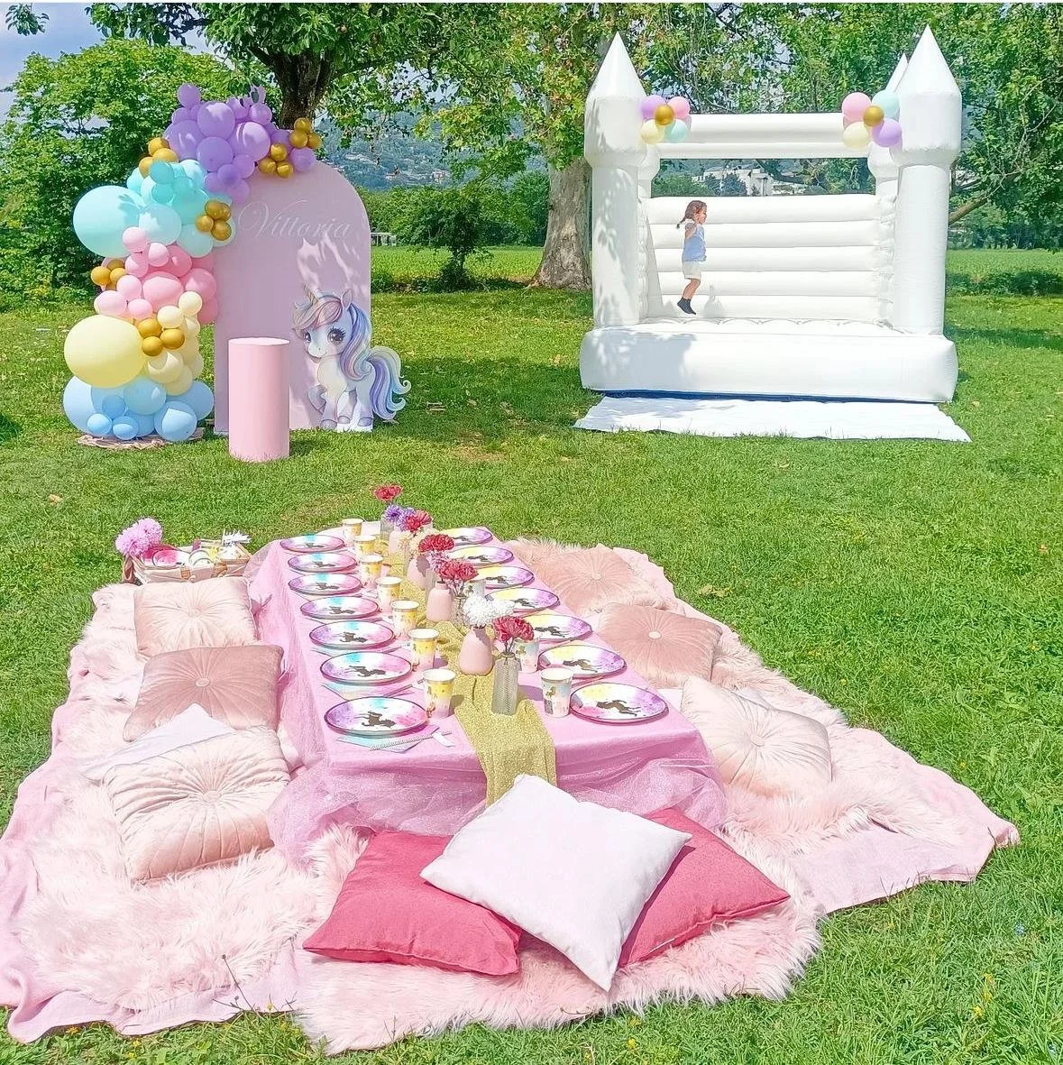 Decorazione per festa bambina con tema unicorno, minimo tavolo rosa coperto, piatti e bicchieri con pattern unicorno, cuscini rosa e bianchi, tappeto rosa, gonfiabile a forma di castello bianco con palloncini decorativi, sfondo di alberi in un parco.