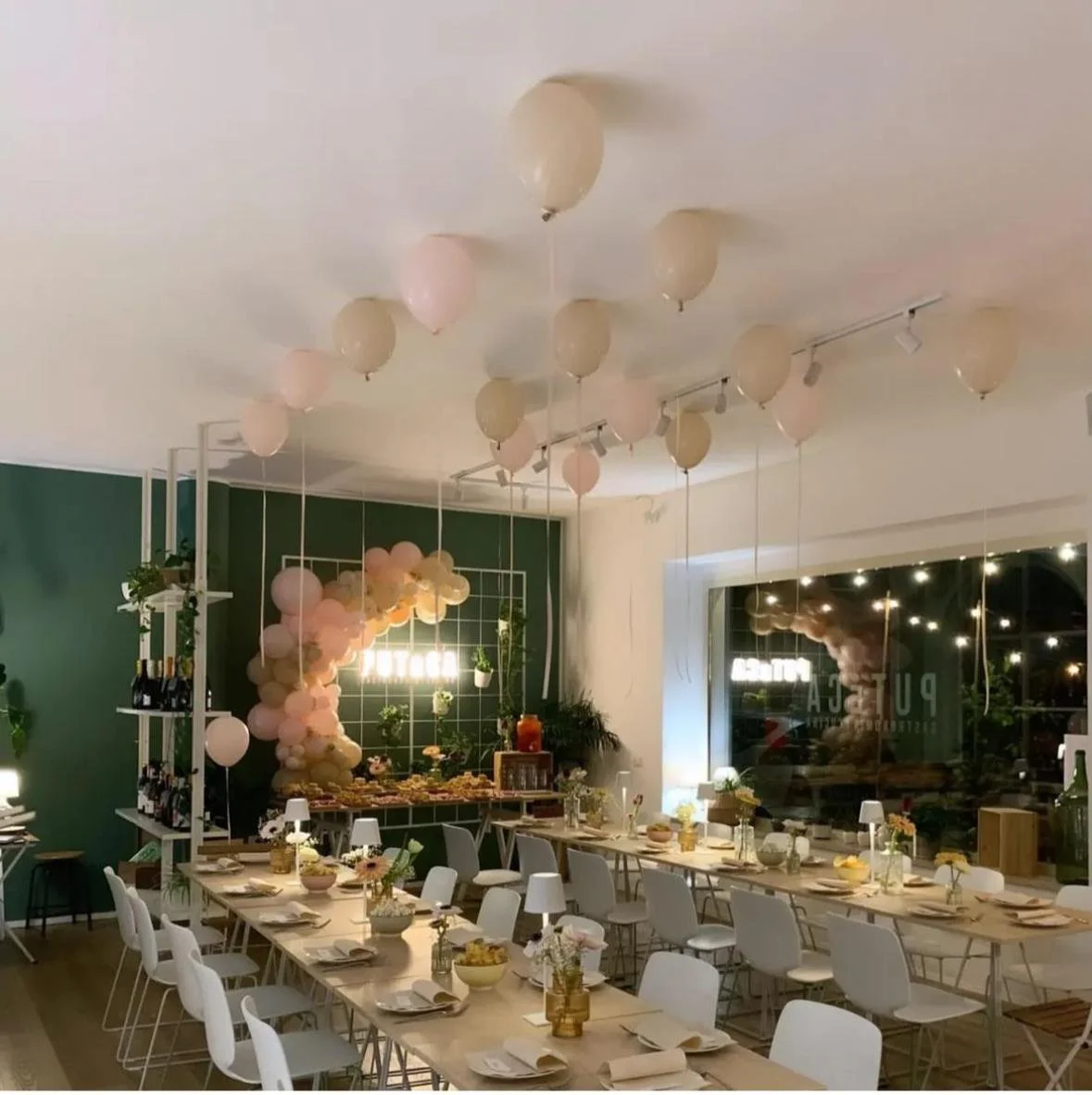 Decorazione di festa con palloncini rosa e crema appesi al soffitto, tavolo apparecchiato per un evento con fiori e cibo, ambientazione elegante e luminosa.