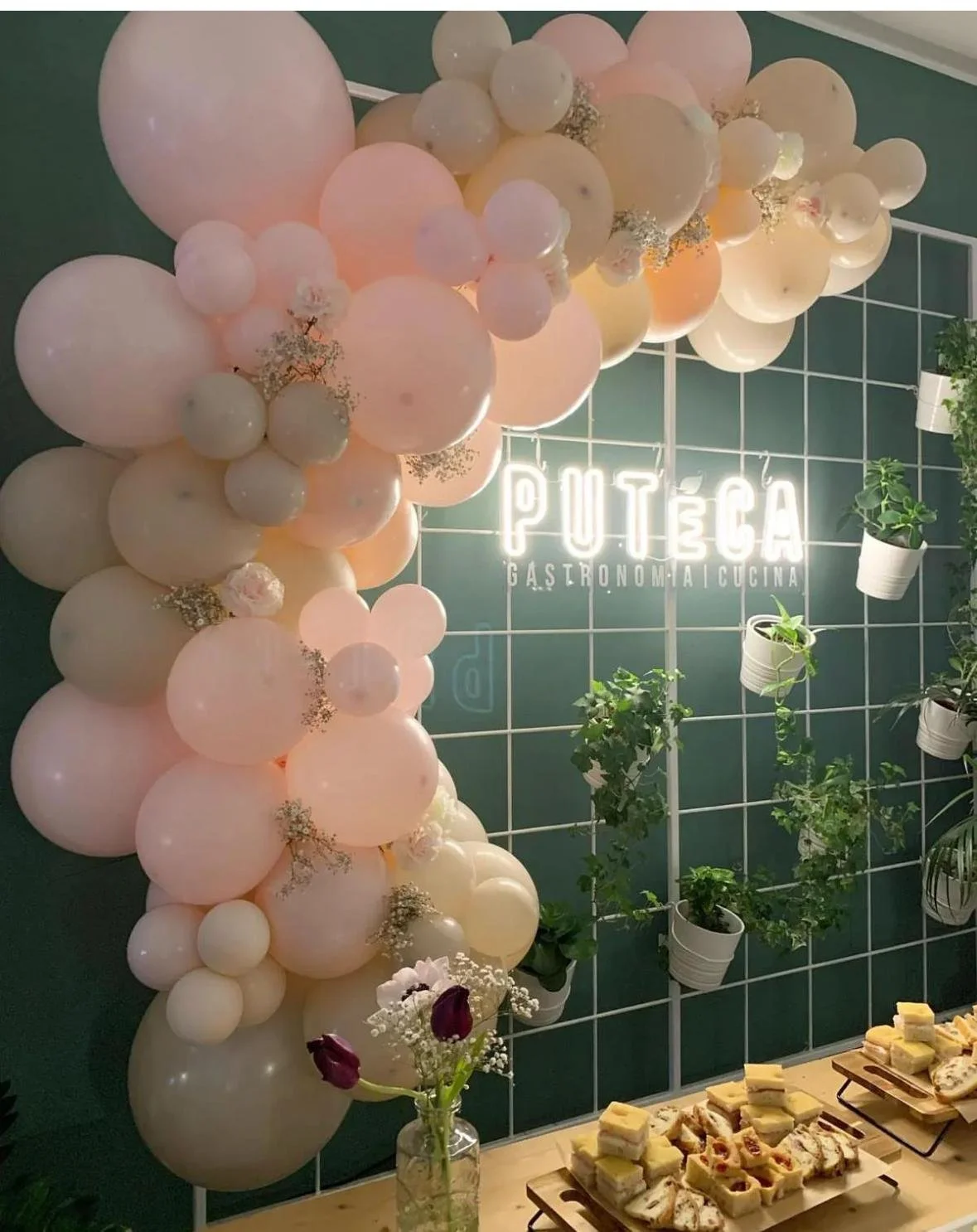 Decorazione con palloncini rosa e bianco, un neon con la scritta 'PUTÉCA GASTRONOMIA CUCINA', piantine in vasi bianchi appese alla parete verde, vassoio con dolci e ciambelle.