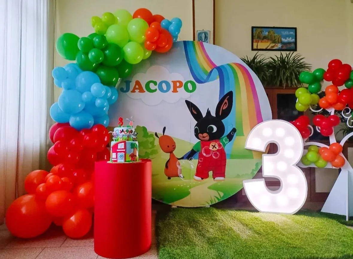 Decorazione di compleanno con palloncini colorati e un grande cartellone con il nome Jacopo e un personaggio animato di un coniglio nero e una lumaca, una torta nera, una grande illuminata con il numero 3.