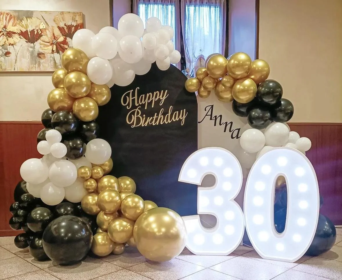 Decorazioni di compleanno con palloncini bianchi, neri e dorati, lettere luminose con il numero 30, e un cartellone nero con scritto "Happy Birthday" e "Anna".