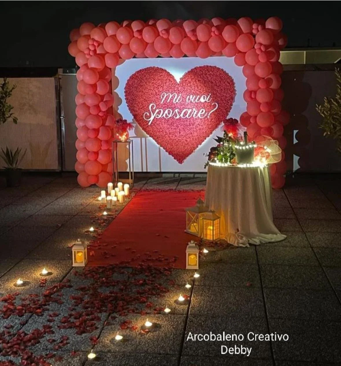 Decorazione romantica per proposta di matrimonio con grande cuore di fiori rossi sullo sfondo, palloncini rosa a formare un arco, resti di petali rossi sul tappeto rosso, candele accese e lanterne, tavolo con fiori e luci. Testo sullo sfondo: 'Mi vuoi sposare?'