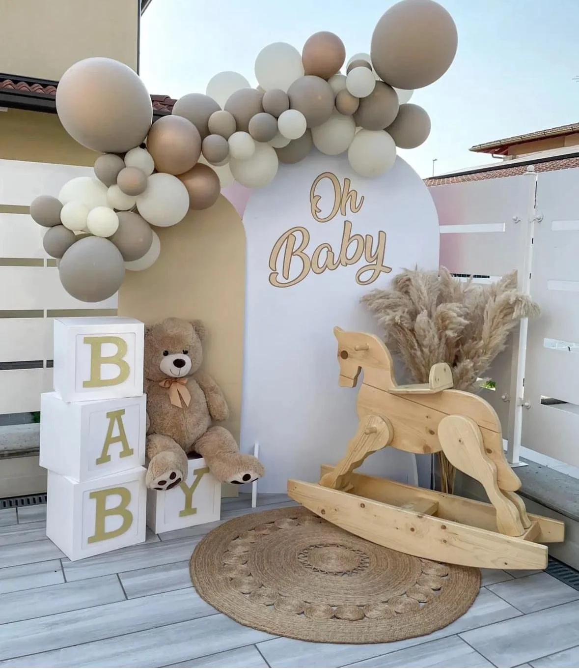 Decorazione per festa di benvenuto baby con palloncini, teddy bear, arcobaleno di pampas, scatole con lettere B, A, B, Y, e uno sgabello a dondolo in legno.