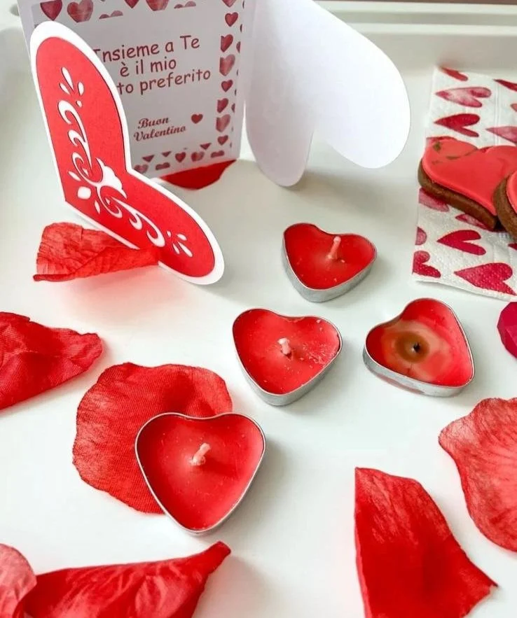 Cuore di cioccolato con candela rossa, petali di rosa, biglietto di San Valentino con cuori rossi e scritto in italiano e un pacchetto decorato a tema cuori.