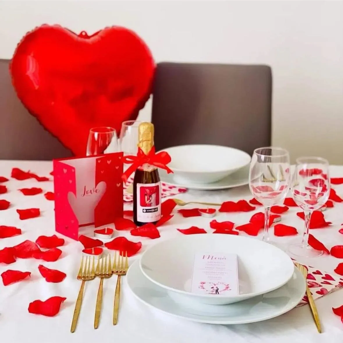 Tavola apparecchiata con decorazioni rosse e romantiche, include un cuore gonfiabile rosso, petali di rosa, una bottiglia di spumante con fiocco rosso, due calici di vino, e un menu, ideale per una celebrazione romantica.