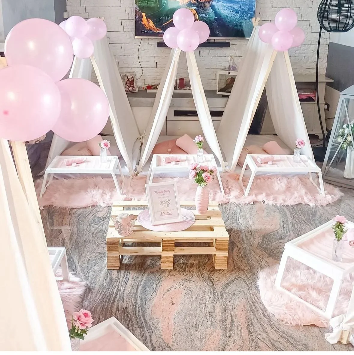 Decorazione di festa con tende, palloncini rosa, tavolini, vasi di fiori rosa, coperte in peluche rosa, e un discorso centrale con fiori. Atmosfera elegante per evento speciale.