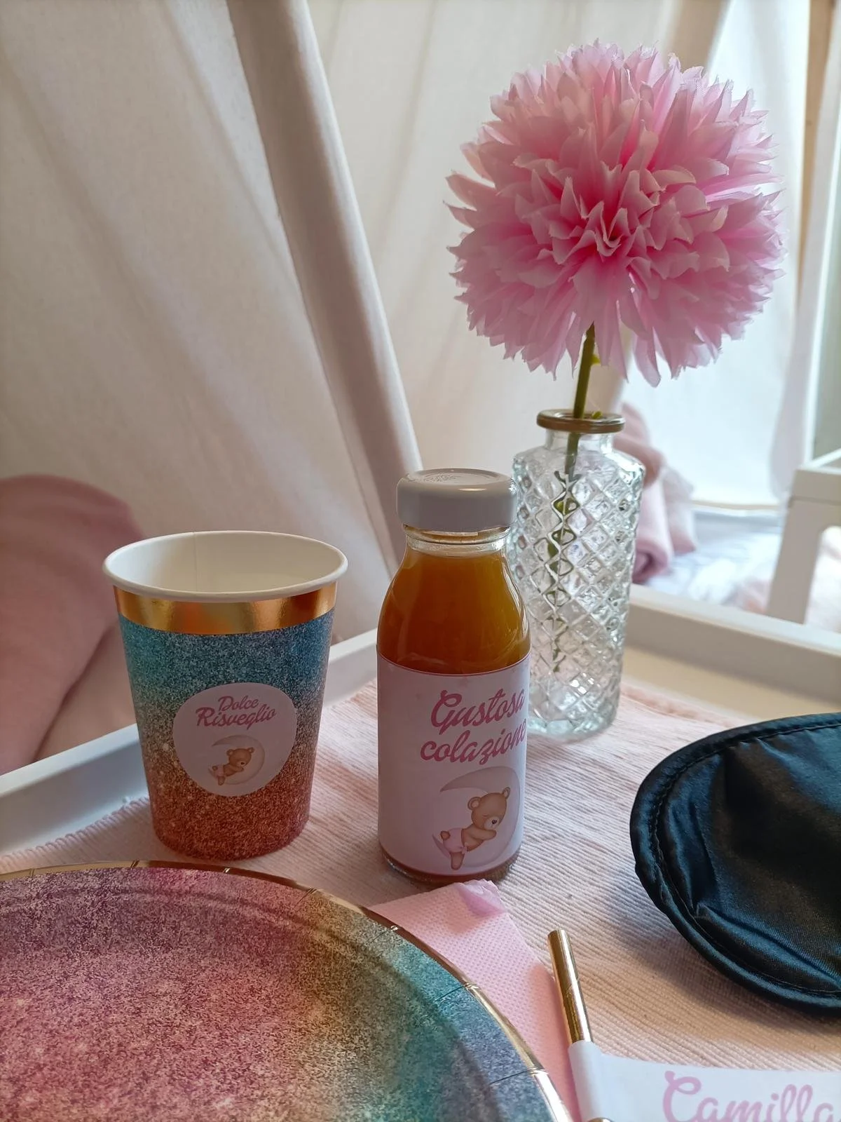 Fiala di vetro con fiore rosa, bottiglia di succo con etichetta rosa e immagine di elefante, e tovagliolo con scritta in italiano, ambientato su tavola con piatto colorato.
