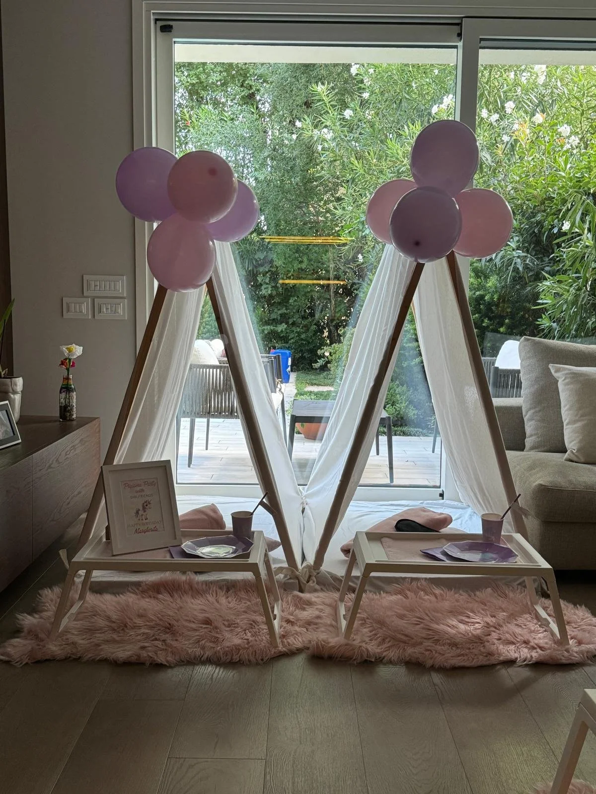 Decorazione festosa con due tende a teepee con palloncini rosa e viola, due tavolini con tovagliette, bicchieri di carta e cane in tessuto, sotto un soffice tappeto rosa, in salotto vicino a un grande finestrone con vista sul giardino.