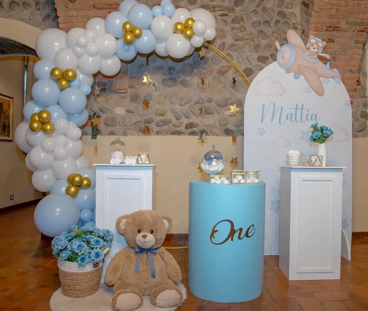 Decorazione per festa di compleanno con balloon bianchi e dorati, un grande orsetto di peluche, una scatola con palline, decorazioni a forma di nuvole e stella in oro, un cartellone con il nome 'Mattia', un sostegno azzurro con la parola 'One', e una