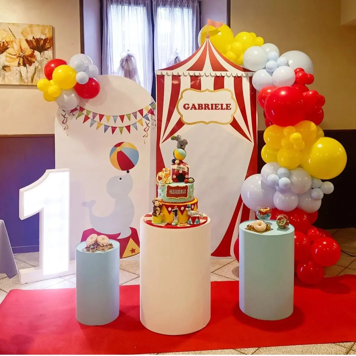 Decorazione di festa di compleanno con tema circus, con un grande dolce, palloncini colorati in rosso, giallo e bianco, e il nome Gabriele scritto in un cartellone circense.