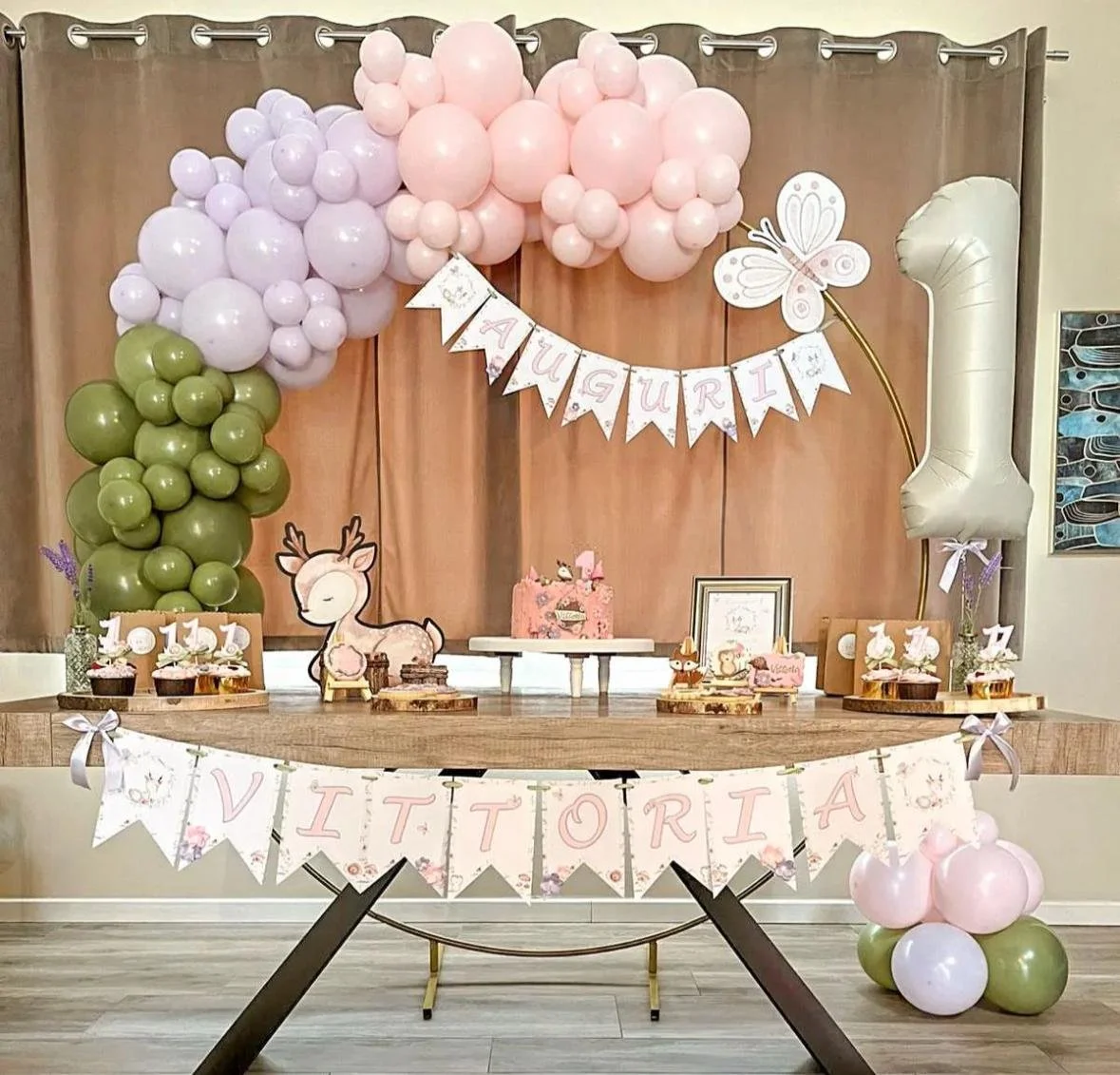 Decorazione di festa con palloncini rosa, lilla, verdi, e decorazioni a tema cervo e farfalla, con dolci, torte, e una ghirlanda con scritto 'Vittoria' e 'Auguri'.